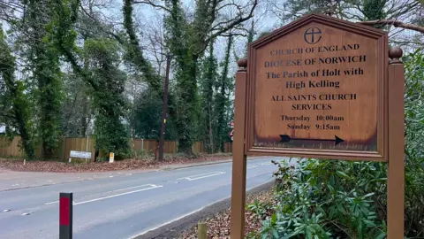 A wooden church sign stands next to a road. Trees line the road on the left hand side. A road sign into "Avenue Road" can also be seen on the opposite side of the road.