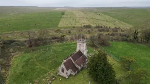 An aerial image of the old church on a misty overcast day. It is a light stone structure with a tall tower at the back and an arched window at the front. It is in the middle of a large grassy plain with trees dotted around it. There are a few gravestones visible around it.