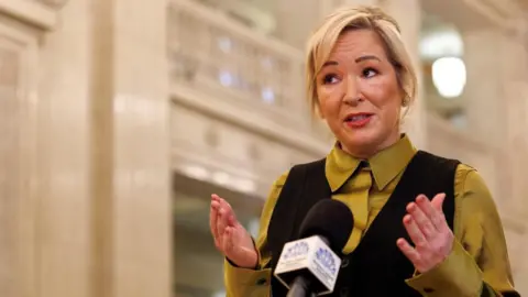 First Minister Michelle O'Neill standing in Stormont's Great Hall in front of a microphone and speaking. She has her hands raised in an expressive manner. She's wearing a yellow-green top with a black waistcoat.