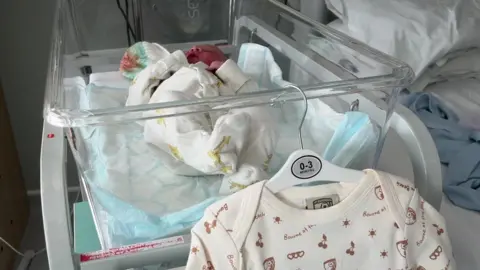 A baby lies in a clear hospital crib. A white sleepsuit with official AFC Bournemouth branding hangs from a coat hanger at the end of the crib. 