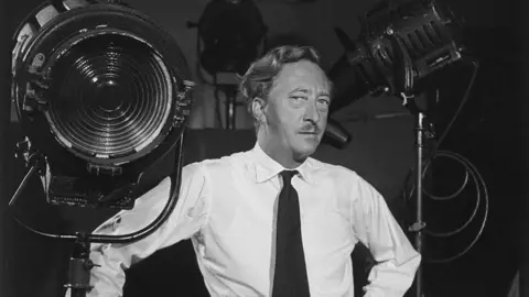 Baron/Getty Black and white image of Walter Greenwood wearing a black tie and white shirt with his arm resting on a stage light and standing in front of other lights.