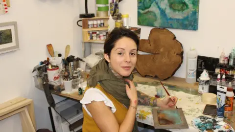 Andreea, smiling to camera, at her work table, with a paintbrush in her hand and a painting in process. Artist equipment such as paints and brushes surround her