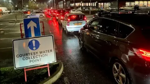 Cars queuing for a water collection point. There is a sign that points into its direction. It is night time.