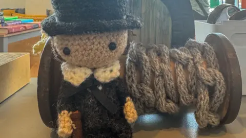 Crochet man in a Victorian outfit with top hat and a crocheted reel of chain behind.