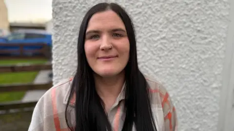 Stacey Smyth has long dark hair. She has a hoop piercing on the right side of her nose. She's wearing an orange and beige checked top. A house is behind her. 