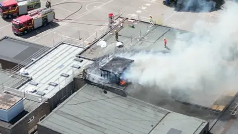 An aerial photograph taken above Ormiston Cliff Park Primary  Academy in Gorleston. Smoke is billowing out from the roof of the building. There are fire fighters on the roof trying to extinguish the flames.