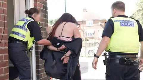 Northumbria Police Two police officers, one on each side of Leanne Hodgson, who has a black top slipping off her shoulders and is wearing handcuffs, as they arrest her at her home address 