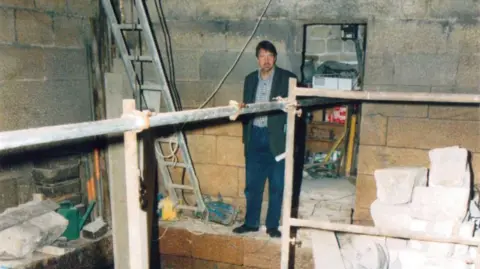 Hebden Bridge Mill A man in a blazer, shirt and jeans stands in a building site 