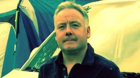 Man - William Horne - wearing a blue open-necked shirt and smiling at the camera with tents behind him.