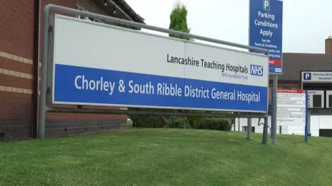 A blue and white sign saying Chorley and South Ribble District General Hospital in white on a blue background below a white bar that says Lancashire Teaching Hospitals NHS Foundation Trust. It sits on a grassed area by a red brick hospital building