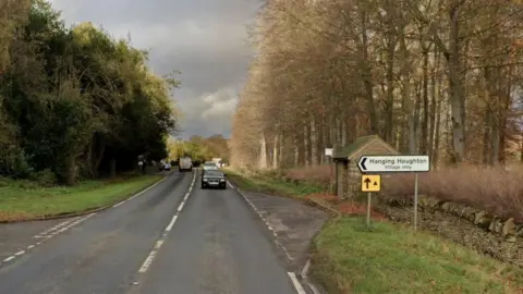 Single carriageway rural road with grass either side.  There is a sign to "Hanging Houghton - village only) in the foreground, in front of a stone bus shelter. There is a turning off to the right. There are tall trees beyond the grass on either side of the road. Two cars are heading towards the camera, with a van travelling in the opposite direction.
