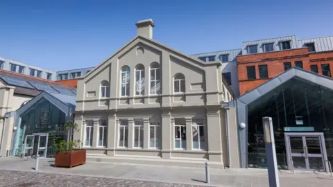 Getty Images The Titanic Hotel, formerly the drawing offices of Harland & Wolff. In the centre is an older building painted a dark cream, flanked by modern glass and metal extensions with pointed roofs. Behind it red brick buildings can be seen.