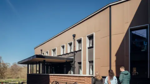 Jim Stephenson A mainly light brown building has two visible storeys. Three people are on the bottom right of the photo. Trees and grass are in the distance on the left. 
