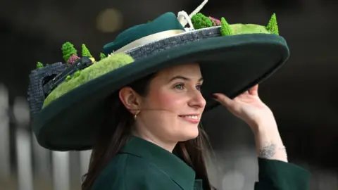 A woman with long black hair and a dark green coat wears a large-brimmed hat decorated around the rim with a train track. An engine is emerging from a tunnel on the hat.