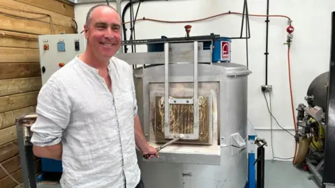 A smiling, balding man, wearing a white shirt, stands in front of a glass-shaping furnace, with one hand on the door handle. 