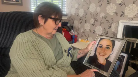 John Fairhall/BBC Sarah Shead is wearing a khaki green cardigan over a darker green top. She is sitting on a chair in her living room and holding a framed picture of her daughter Ms Seaman, who is smiling and wearing a black T-shirt.