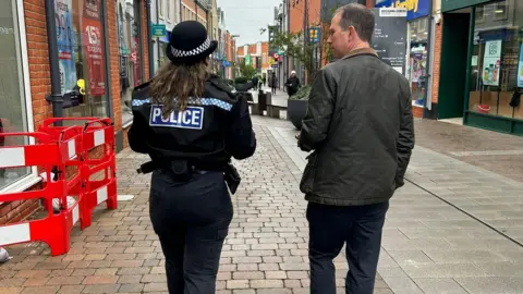  Thames Valley Police and Crime Commissioner PCC Matthew Barber with female police officer in Didcot. They are photographed from the back. They are passing by shops. It is a cloudy day.