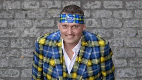 PA Media Doddie Weir is wearing a yellow and blue tartan three piece suit and headband.
