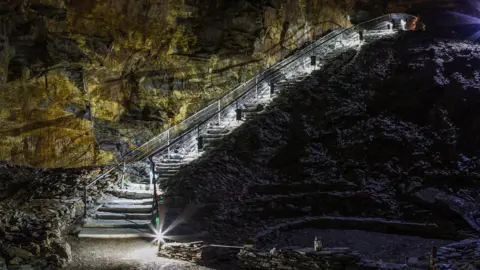 An underground tunnel with steps leading down. It is dark but the steps have white lights on them. The walls are solid rock.