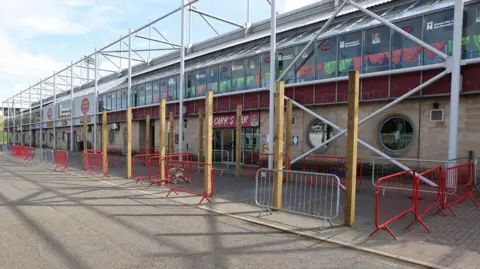 Northampton Town Football Club The outside of a football club, with paving and a road in the foreground. There are metal barriers in white and red dotted around the building.