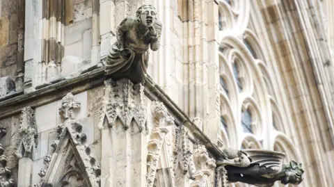 A stone sculpture on the side of York Minster, a large lightly coloured stone cathedral with details around the arches and the windows.