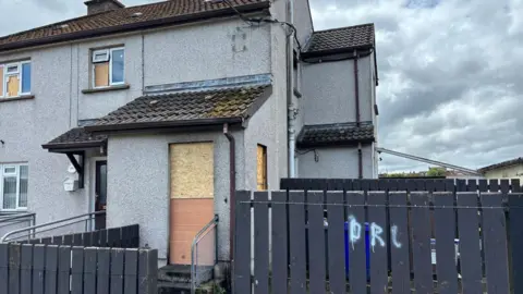A grey end-terrace house with a boarded up door and windows. There is a grey fence in front of the house and a cloudy sky above. 