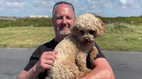 Mark Early stands holding his cockerpoo dog in a car park in Barrow. He is wearing a black t-shirt and black glasses.