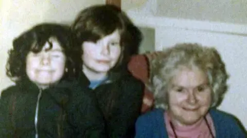 Diane Barnes An old photograph of two young girls with dark hair and their grandmother. The girls are standing together next to an older woman who's sitting in a chair. They're all looking at the camera. 