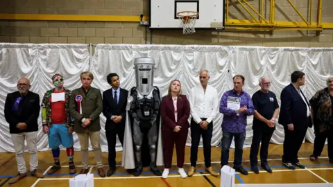 Jack Taylor/Reuters Eleven of the candidates at the 2026 Clacton by-election stand in a line on the floor of a sports hall. Count Binface is prominent in the middle, wearing his dustbin headgear.