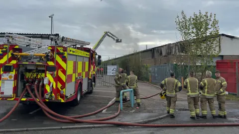 Edd Smith/BBC A fire truck parked outside a commercial building. There are firefighters standing next to it. To the right is a building with smoke damage.