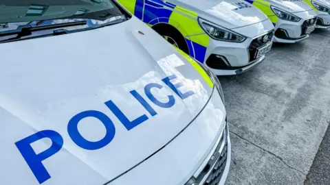 A close-up image shows the bonnet of a police car. "Police" is written in big blue letters. Several other police cars can be seen parked up alongside. 