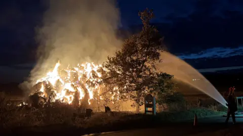 A haystack in a field on fire. The photo has been taken at night however, the haystack has been illuminated by orange flames. Smoke is also billowing from the stack and into the sky. A firefighter, with a hose, is standing on the road to try to extinguish the fire.