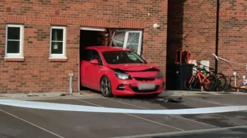 A red car is sticking out of the front of one of a house.