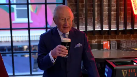 King Charles in a suit holds a pint of Guinness in Guinness Open Gate Brewery, Covent Garden, with a brick wall and colorful window displays in the background.