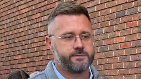 A man with short, light brown hair and brown/grey facial hair. He has glasses and is wearing a blue shirt and grey gilet. He is walking in front of a brown brick wall