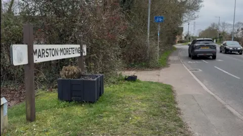 Martin Heath/BBC Road through a village, showing sign spelt "Marston Moreteyne" mounted on a grass verge with a wooden planter beneath. There is a hedge in the background and two cars are passing on the road to the right.