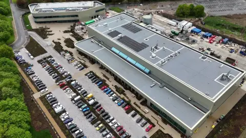A drone shot of a large, long, modern-looking building, and a smaller one next to it. There is a car park with about 100 cars in it.