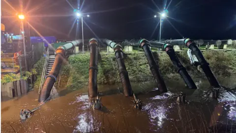 Six large pipes next to an industrial site leading down into a body of water which is dark brown in colour. The whole site is floodlit. 