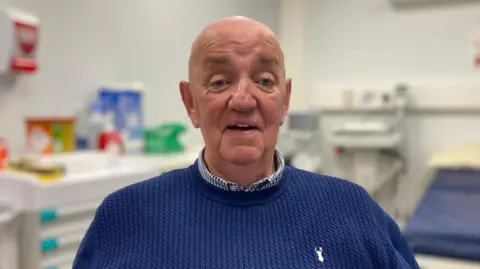 BBC Jim Ross wearing a white and blue checked shirt under a blue jumper. Soft focus in the background is a hospital bed and equipment. 