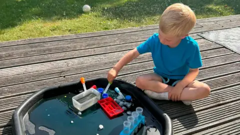 Supplied Reggie sits on some decking outside stirring some mixture is a large plastic container. He is wearing a blue T-shirt and shorts. 