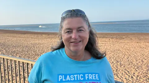 BBC Group leader Jill Gathercole she has shoulder length dark hair and is wearing a blue t shirt. She is stood in front of a long stretch of beach. 