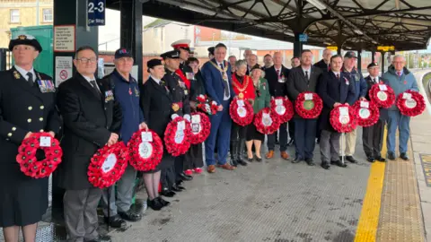 A mixture of men and women, stood side by side, dressed in uniform holding poppy wreaths. They're stood on a train platform. 