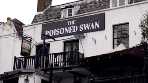 Handout A view from street level of the pub signage. It is a large black and white wall-mounted sign saying 'The Poisoned Swan' in capital letters, on the white stone washed pub building, which has black framed windows and a black painted balcony.