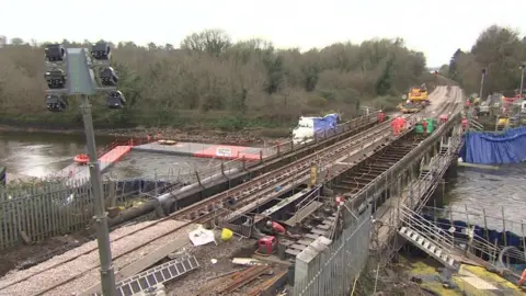 Looking down on the bridge from a pedestrian footbridge, several workers can be seen in orange high visibility overalls and hard hats. Temporary floodlights can be seen in the foreground, while to the left of the bridge a grey and orange pontoon floats on the water. An orange plastic dingy sits on top of it.