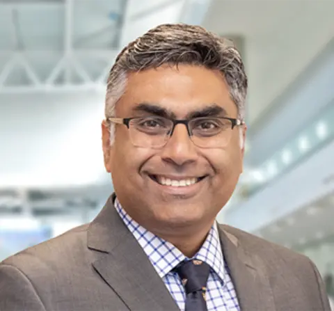 Vikas Khanduja vestindo um blazer, camisa xadrez azul e branca e uma gravata azul. Ele também usa óculos de armação quadrada e está sorrindo para a câmera. 