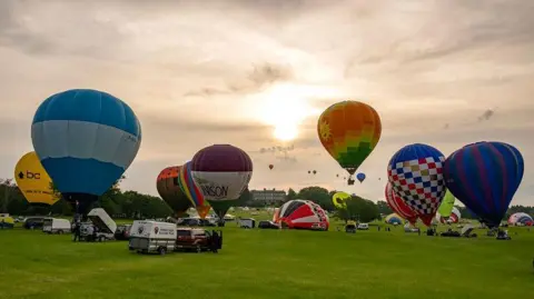 A number of hot-air balloons take off in a field in the grounds of an estate. Several vehicles are parked underneath the balloons. A large hall can be seen in the distance.