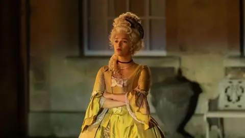 BBC/Bad Wolf Productions/HBO/Simon Ridgway Actress Marisa Abela, as Yasmin in Industry. She is pictured wearing period costume - a yellow ballroom gown  from the 18th century French court, and a blonde wig. She stands with her arms crossed, with the sandstone walls of a stately home behind her.