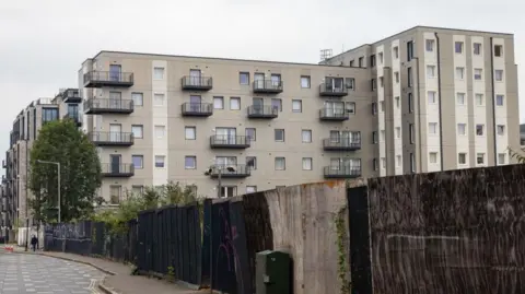 A tall apartment building with grey exterior cladding by a fence and road 