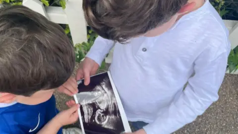 Two children look at a picture of a black-and-white baby scan - one boy wears a blue football shirt and the other a pale grey long-sleeved top 