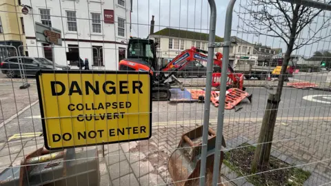 Fencing erected around an orange digger at the site of a hole in a road in Kingsbridge, Devon. It is a busy road with shops along the street. A yellow danger sign telling people not to enter because of a collapsed culvert is on the metal fencing.